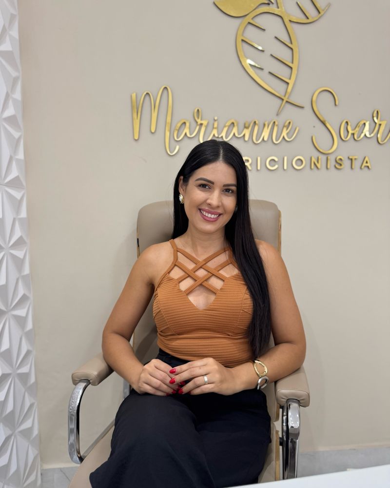 Nutricionista Marianne Soares Machado sentada em frente ao painel dourado com sua logomarca, no consultório